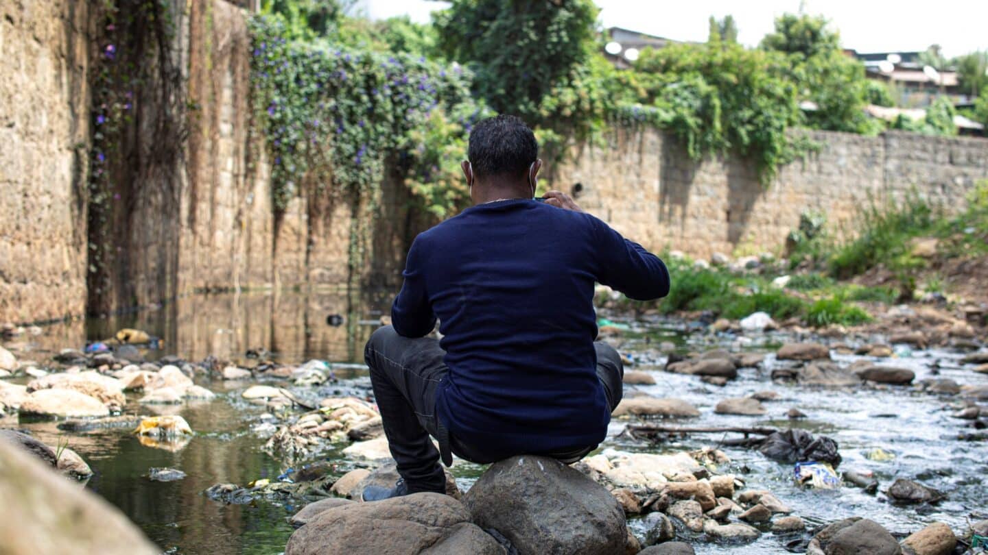 The back of a man facing a small stream with a vegetation-covered brick wall behind the stream.