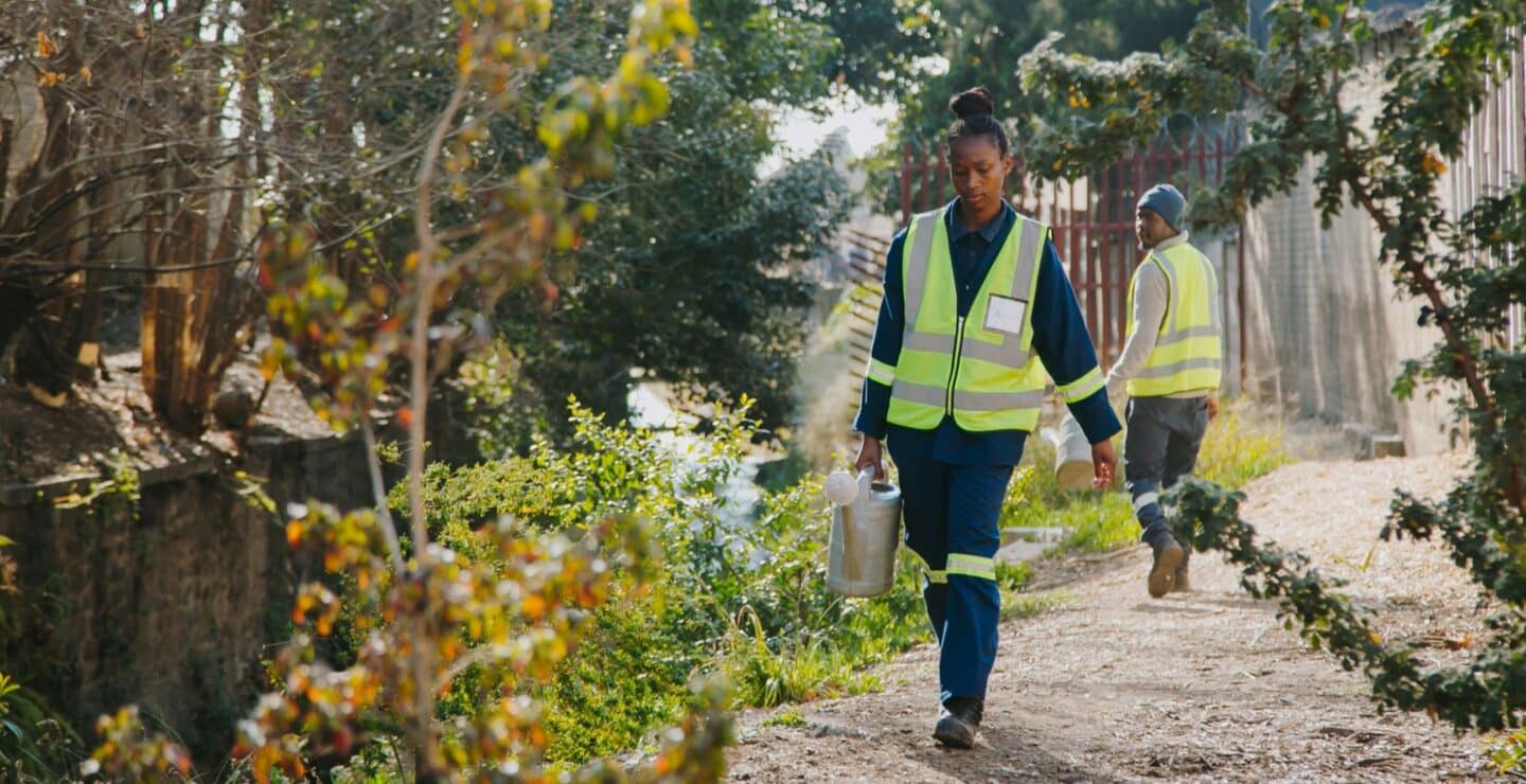 A woman in a construction vest carrying a bucket alongside a river surrounded by trees.